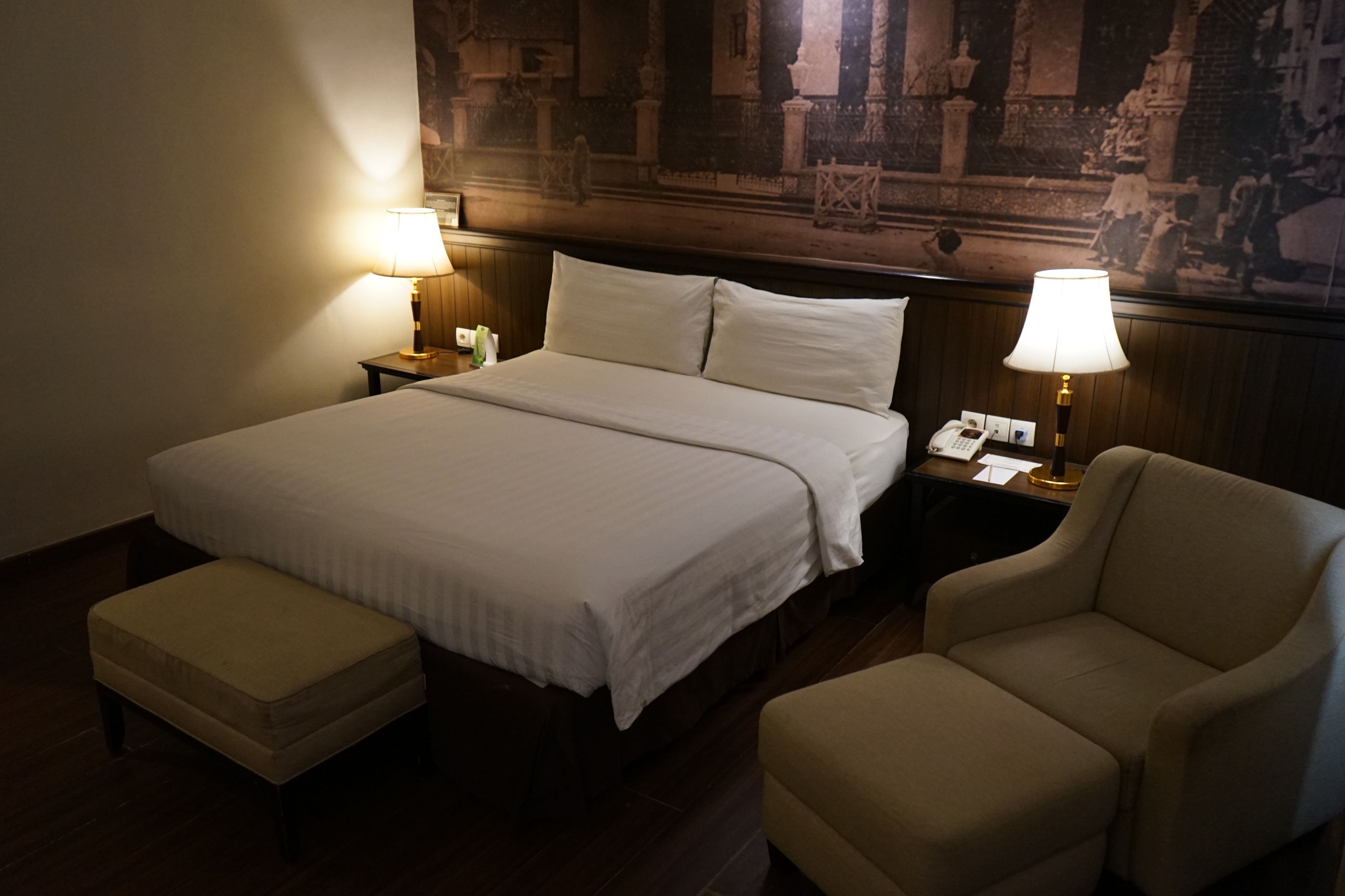 A hotel room with a large bed, two bedside lamps, a telephone, a chair, and a small table.