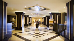 Un'hall dell'hotel con pavimento a scacchi, ampie colonne e un lampadario centrale.