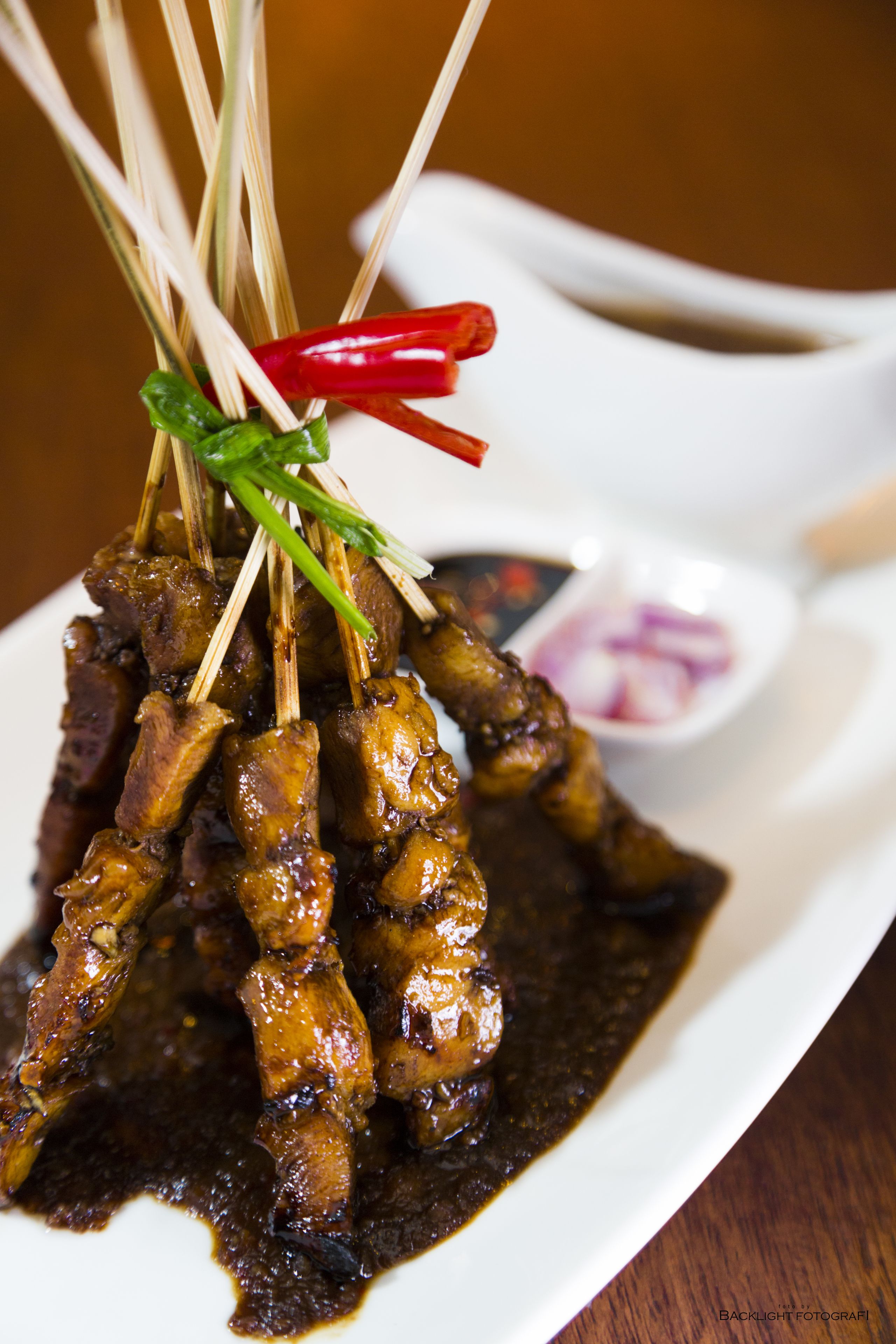 Skewers of meat with a dark sauce, garnished with green onions and red chili peppers, served on a white plate with a side of dipping sauce.
