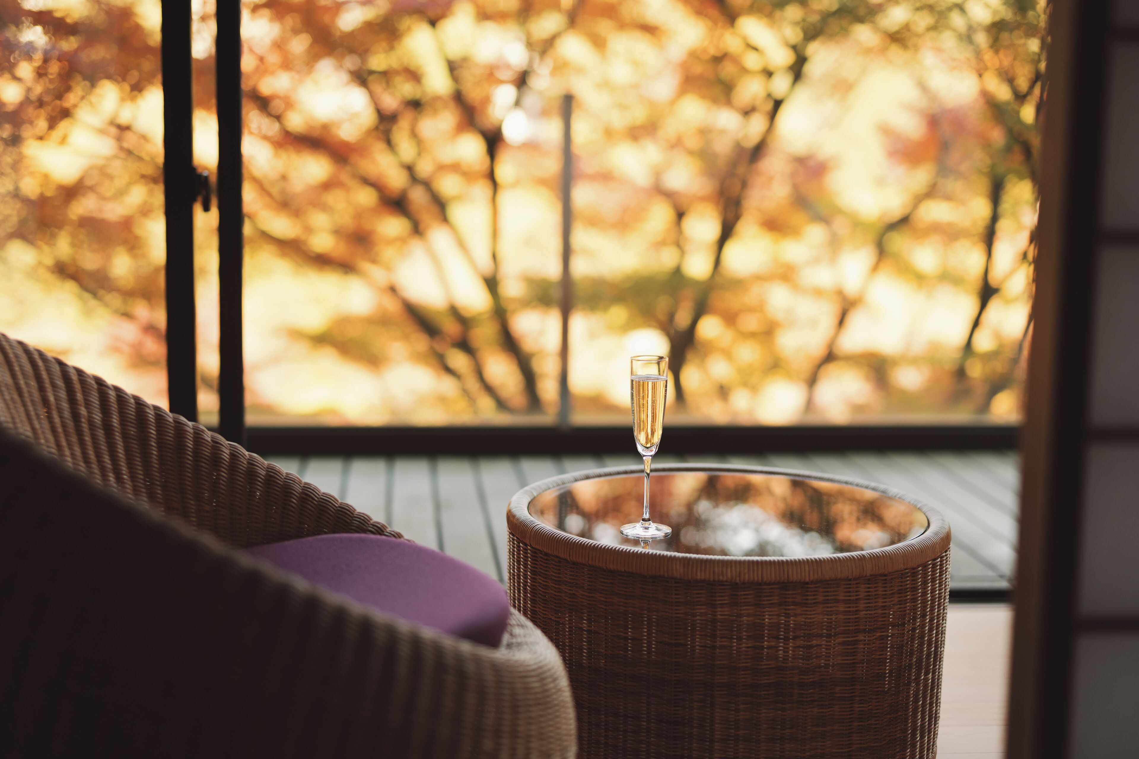 A wicker chair with a cushion, a glass table with a glass on it, and a view of trees through a window.