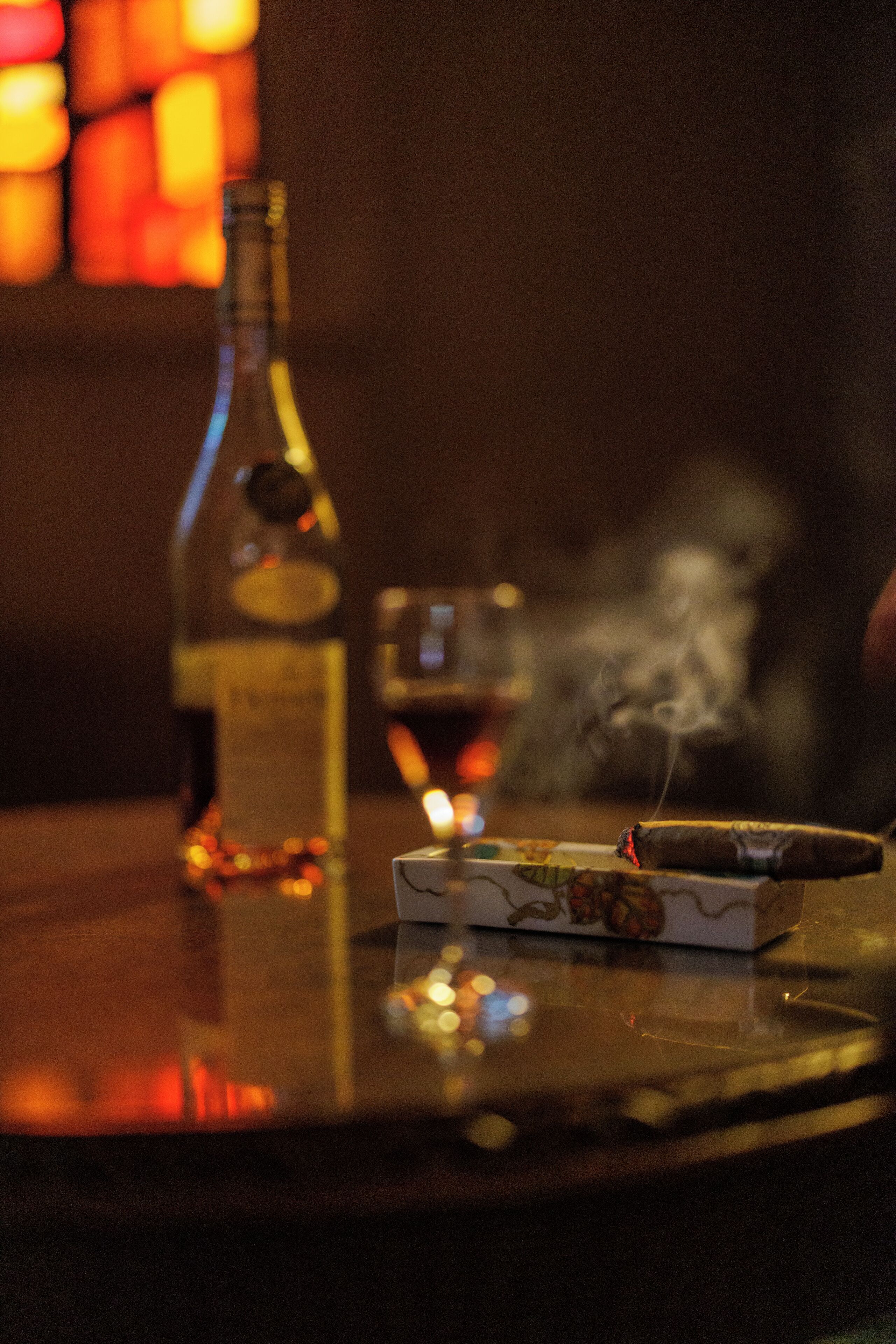 A bottle of liquor, a glass, and a cigar on a reflective surface.