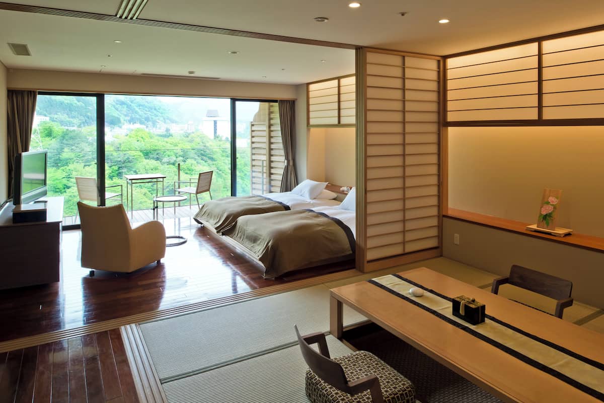 Quarto de hotel com uma grande porta deslizante para uma varanda, uma cama com duas almofadas, uma mesa de cabeceira, uma cadeira e uma pequena mesa com um vaso de flores.