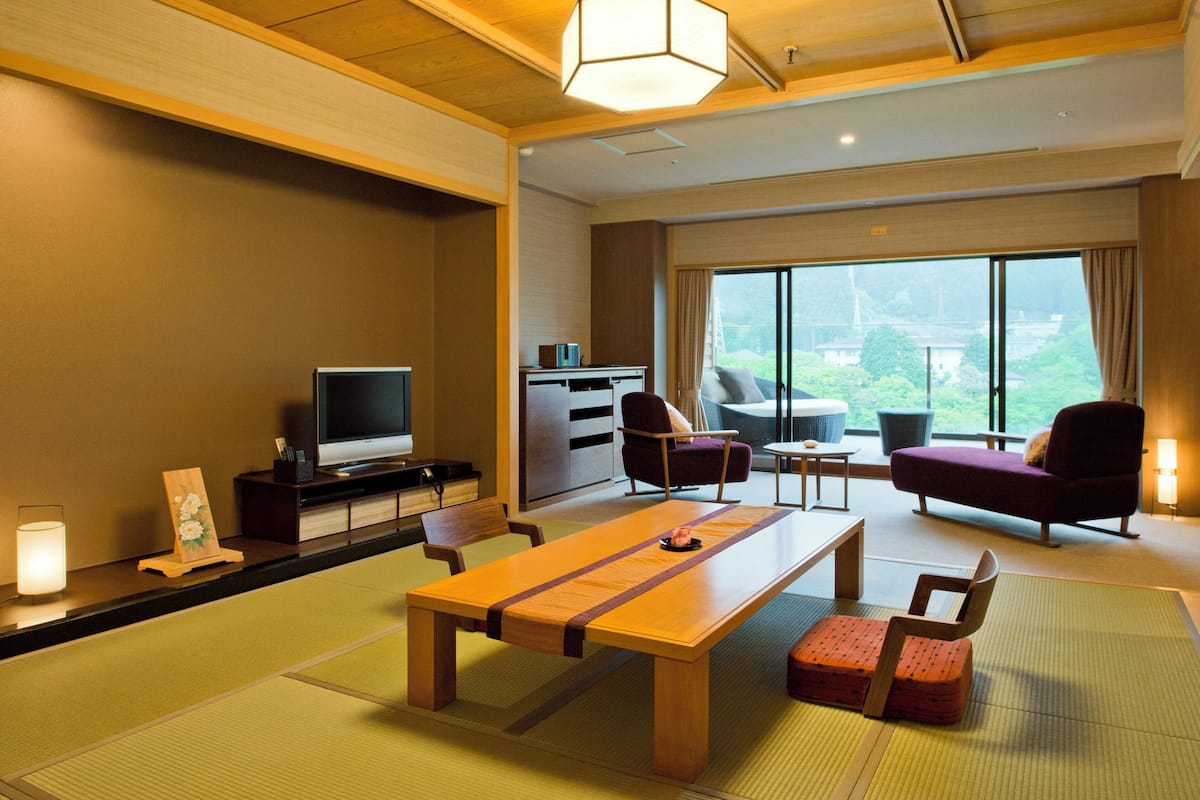Quarto de hotel moderno com piso de madeira, assentos em esteira de tatame, TV de tela plana, escrivaninha e uma grande janela com vista para áreas verdes.