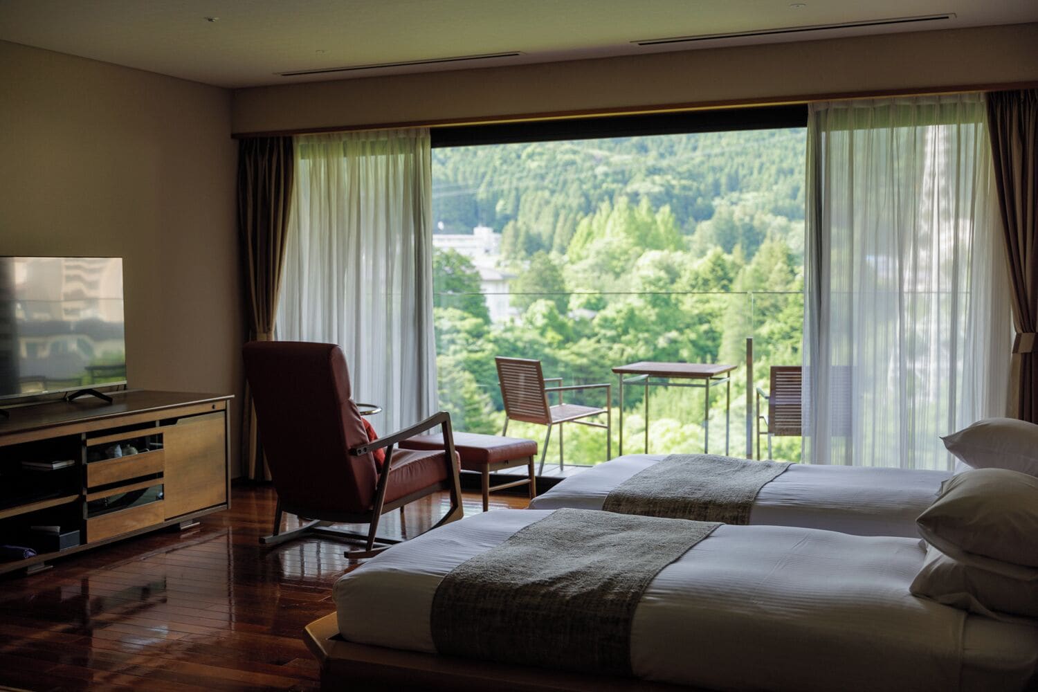 Habitación de hotel con dos camas, televisor, escritorio y balcón con vista a un bosque.