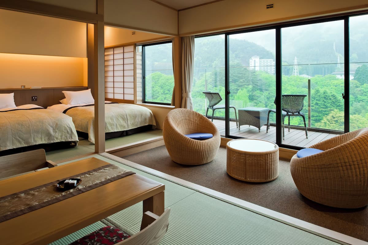 Quarto de hotel com duas camas, varanda com móveis de vime e vista para áreas verdes e edifícios.