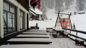 Un área de estar al aire libre cubierta de nieve con bancos y mesas de madera, banderas y un bosque nevado al fondo.