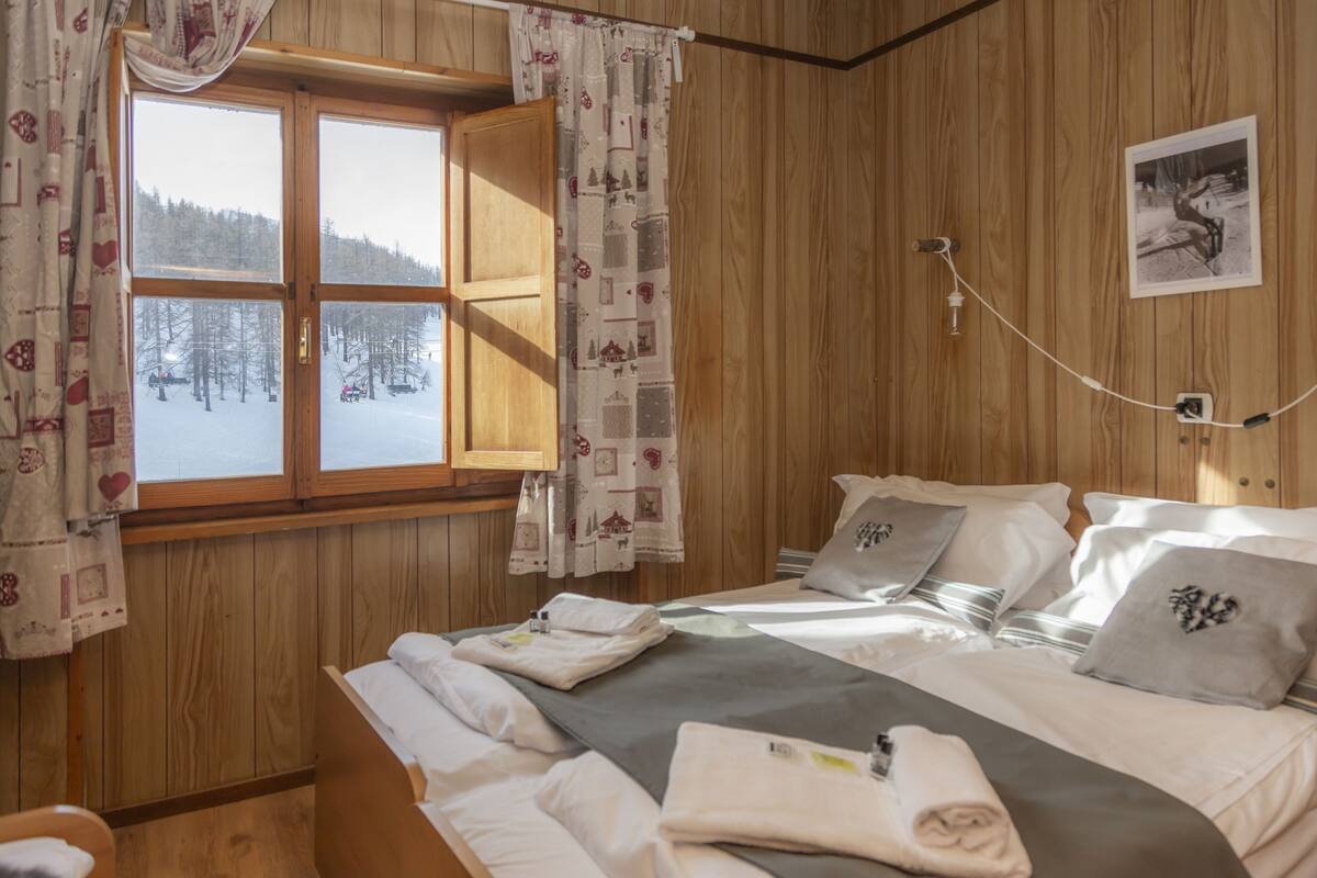 A wooden room with a bed, a window with curtains, a wall-mounted light, and a framed picture.