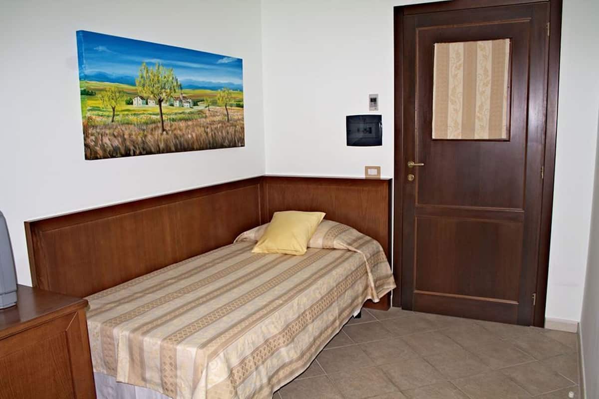 A single bed with a patterned bedspread, a wooden headboard, a door with a decorative panel, and a landscape painting on the wall.