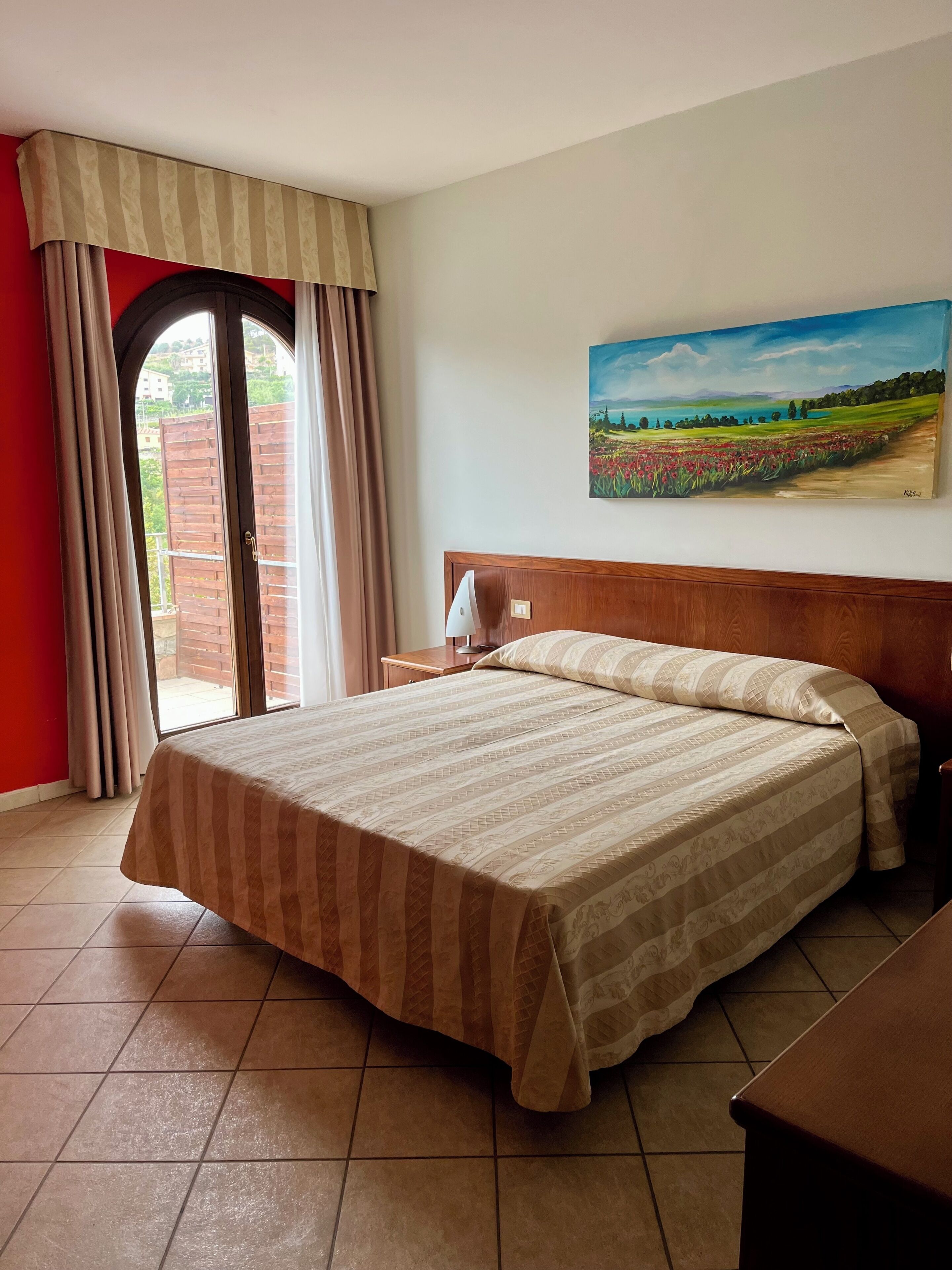 A hotel room with a bed, a nightstand, a painting, and a view of a garden through a window.