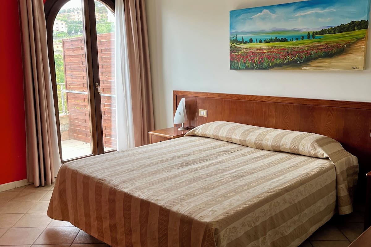 A hotel room with a bed, a nightstand, a painting, and a view of a garden through a window.