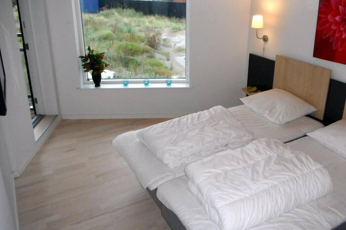 A bedroom with a large bed, a window with a view of greenery, a wall-mounted lamp, and a red flower painting.