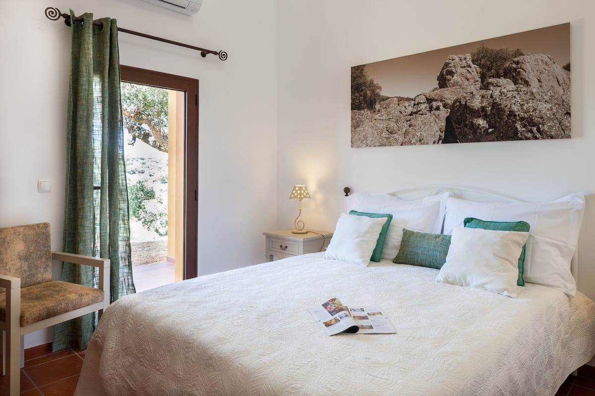 A neatly made bed with white bedding and green pillows, a bedside table with a lamp, a chair, and a large framed picture on the wall.