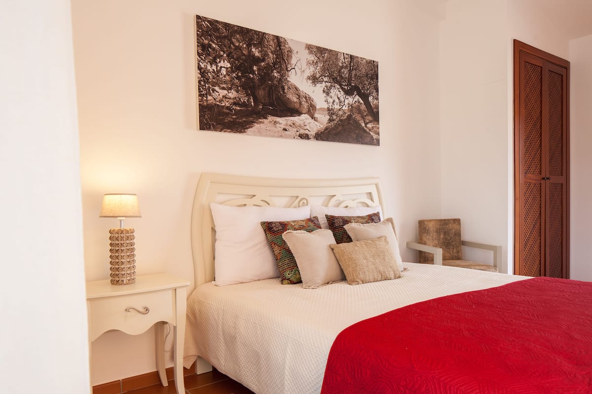 A bedroom with a white bed, a red blanket, a nightstand with a lamp, a chair, and a wooden door.