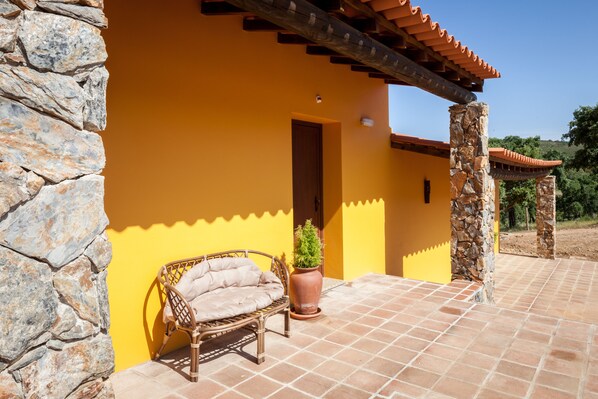 A stone wall, a wicker chair, and a potted plant outside a yellow building with a tiled patio. - Monte da Esperança (Arronches)