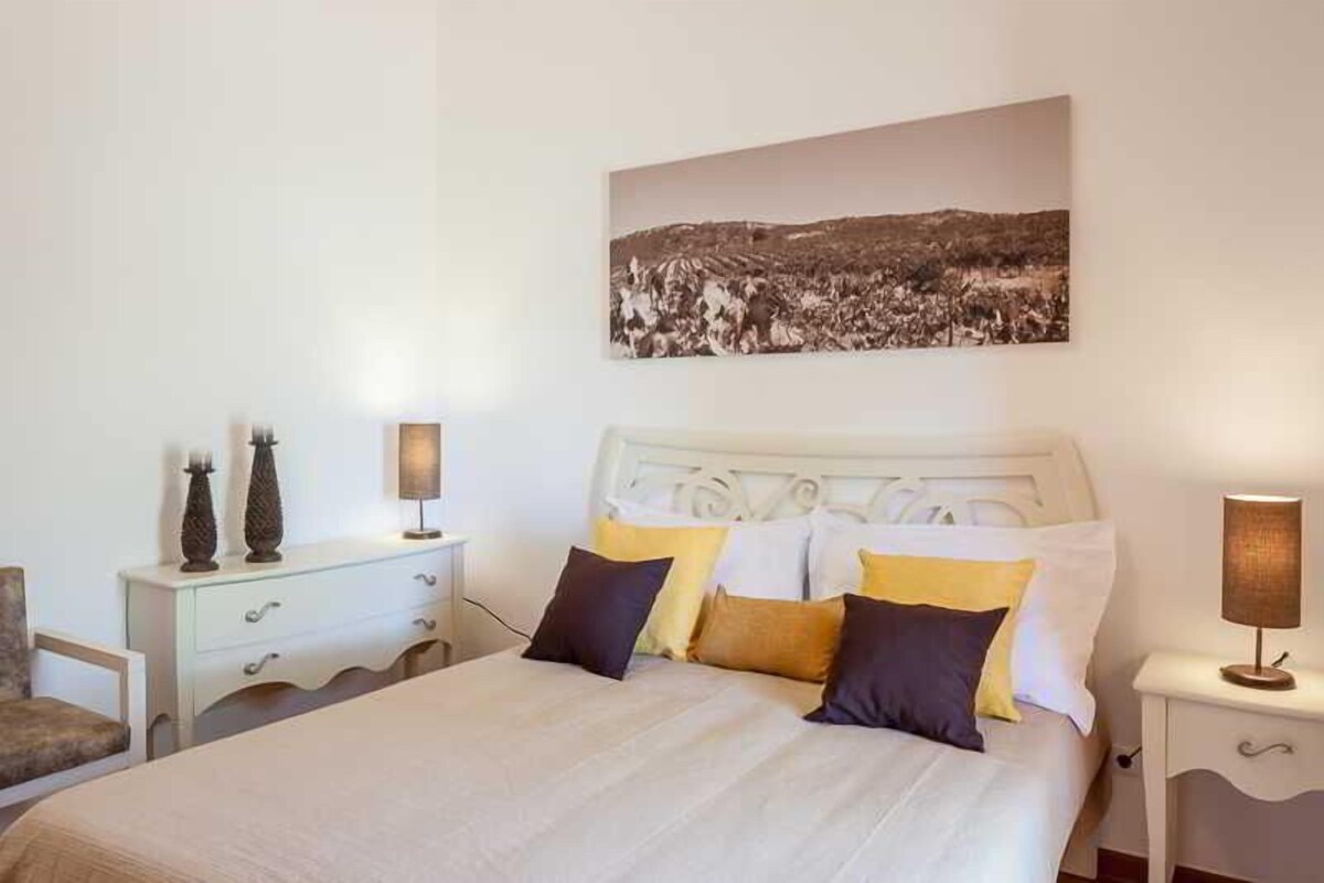 A bedroom with a bed, bedside tables, a nightstand, a chair, and a wall-mounted picture.