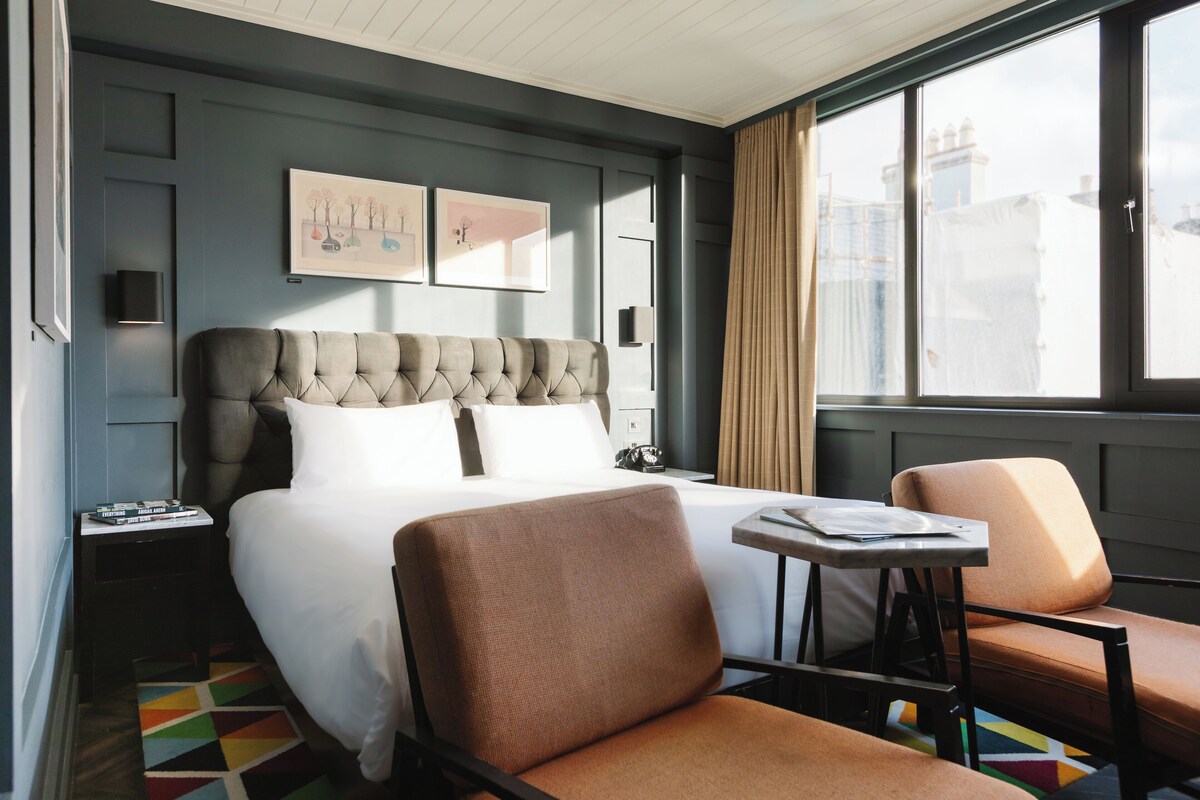 A hotel room with a large bed, a small table, two chairs, a nightstand, and a window with a view of buildings.