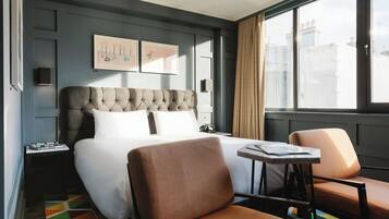 A hotel room with a large bed, a small table, two chairs, a nightstand, and a window with a view of buildings.