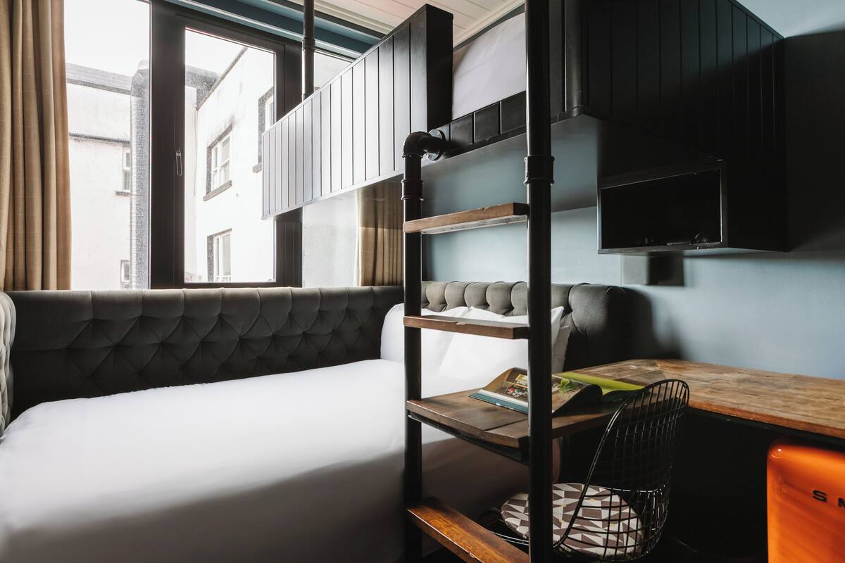 A compact room with a bunk bed, a desk, and a chair. There is a window providing natural light, and a television mounted on the wall.