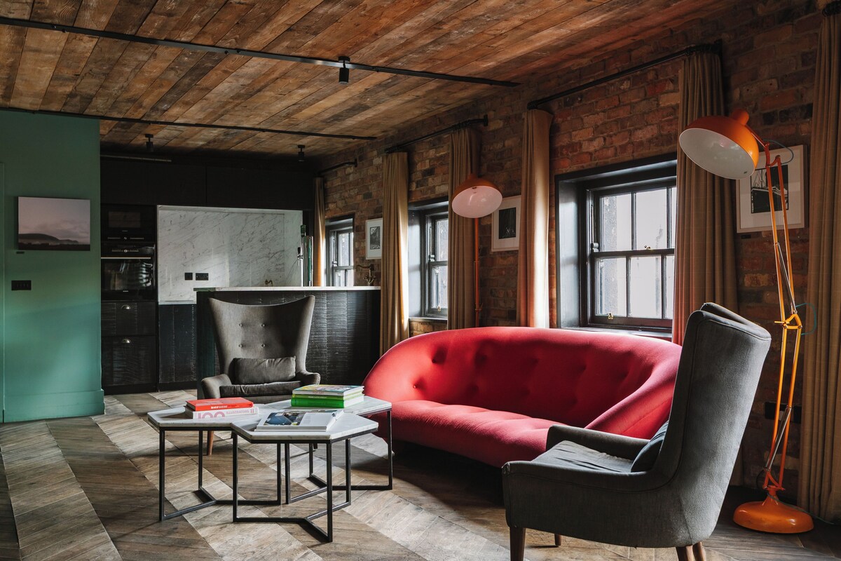 A modern living room with a red sofa, a grey armchair, and a coffee table. There is a brick wall and a window with curtains.