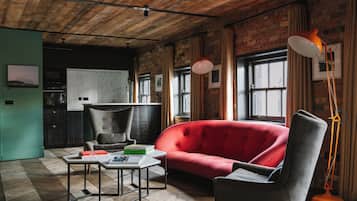 Una sala de estar moderna con un sofá rojo, un sillón gris y una mesita de centro. Se observa una pared de ladrillo y una ventana con cortinas.