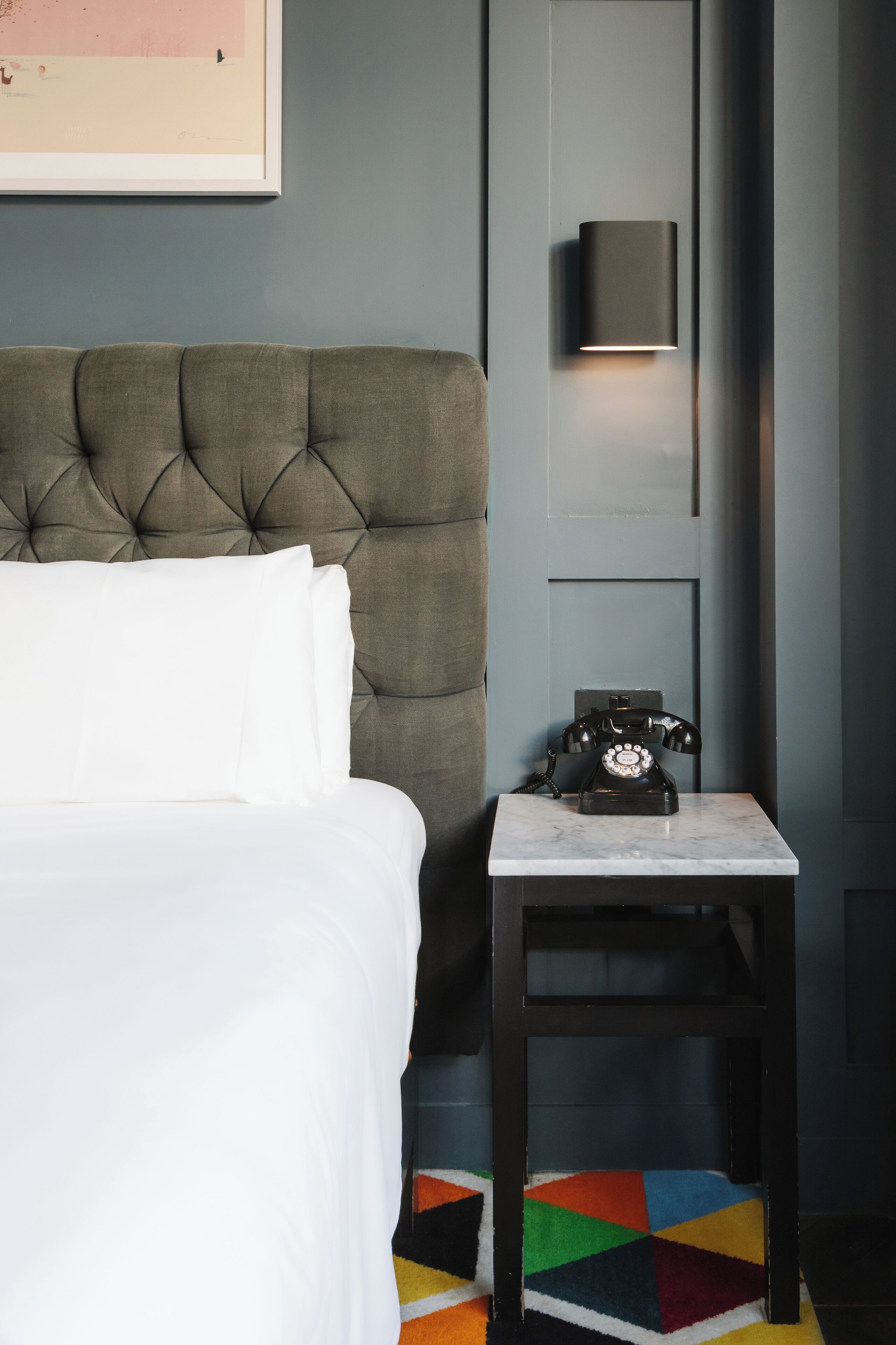 A bed with white linens, a tufted headboard, a black telephone on a marble table, and a colorful geometric rug.