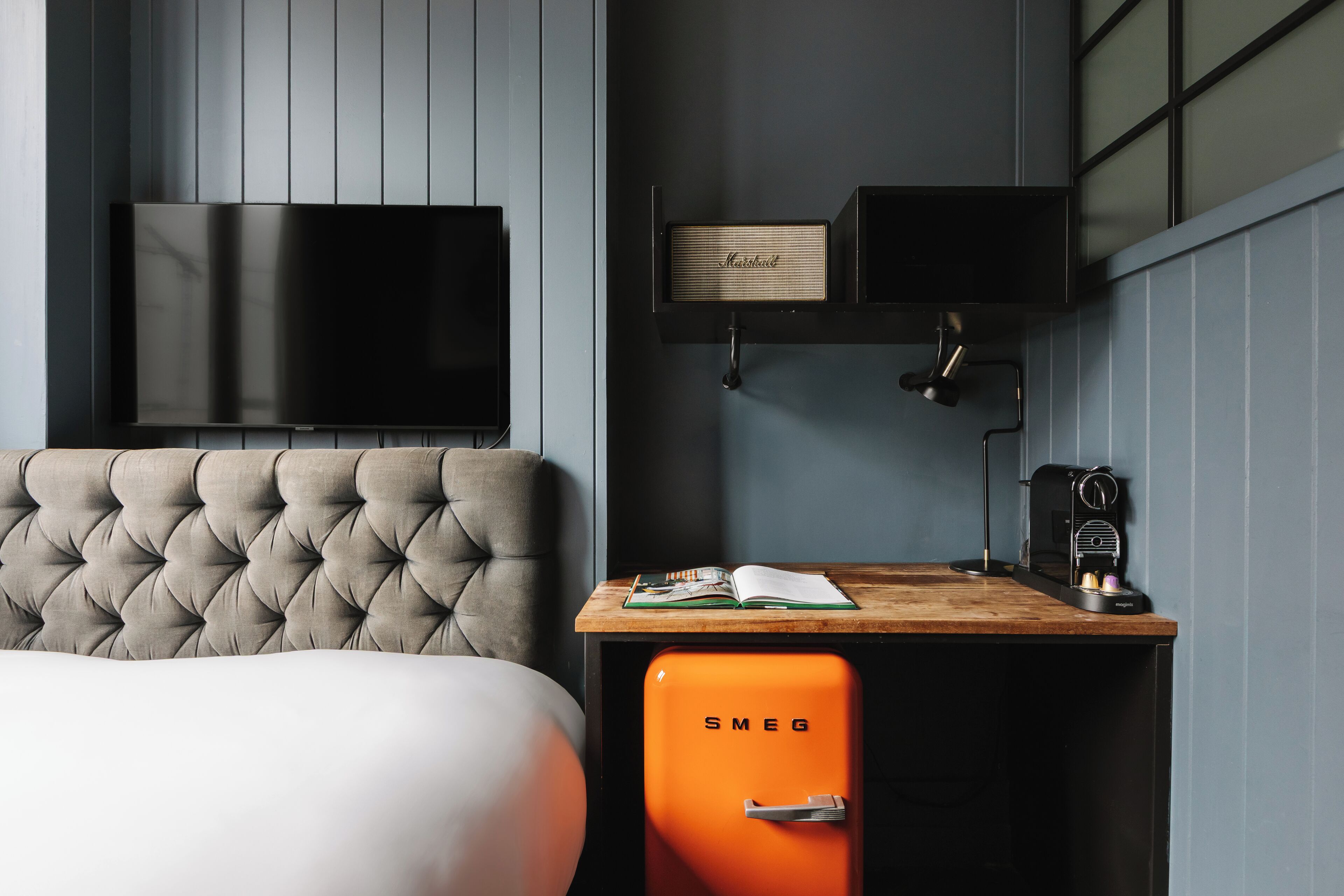 A modern room with a tufted headboard bed, a flat-screen TV mounted on the wall, a wooden desk with a book, a vintage orange Smeg coffee machine, and a Marshall speaker on a shelf.