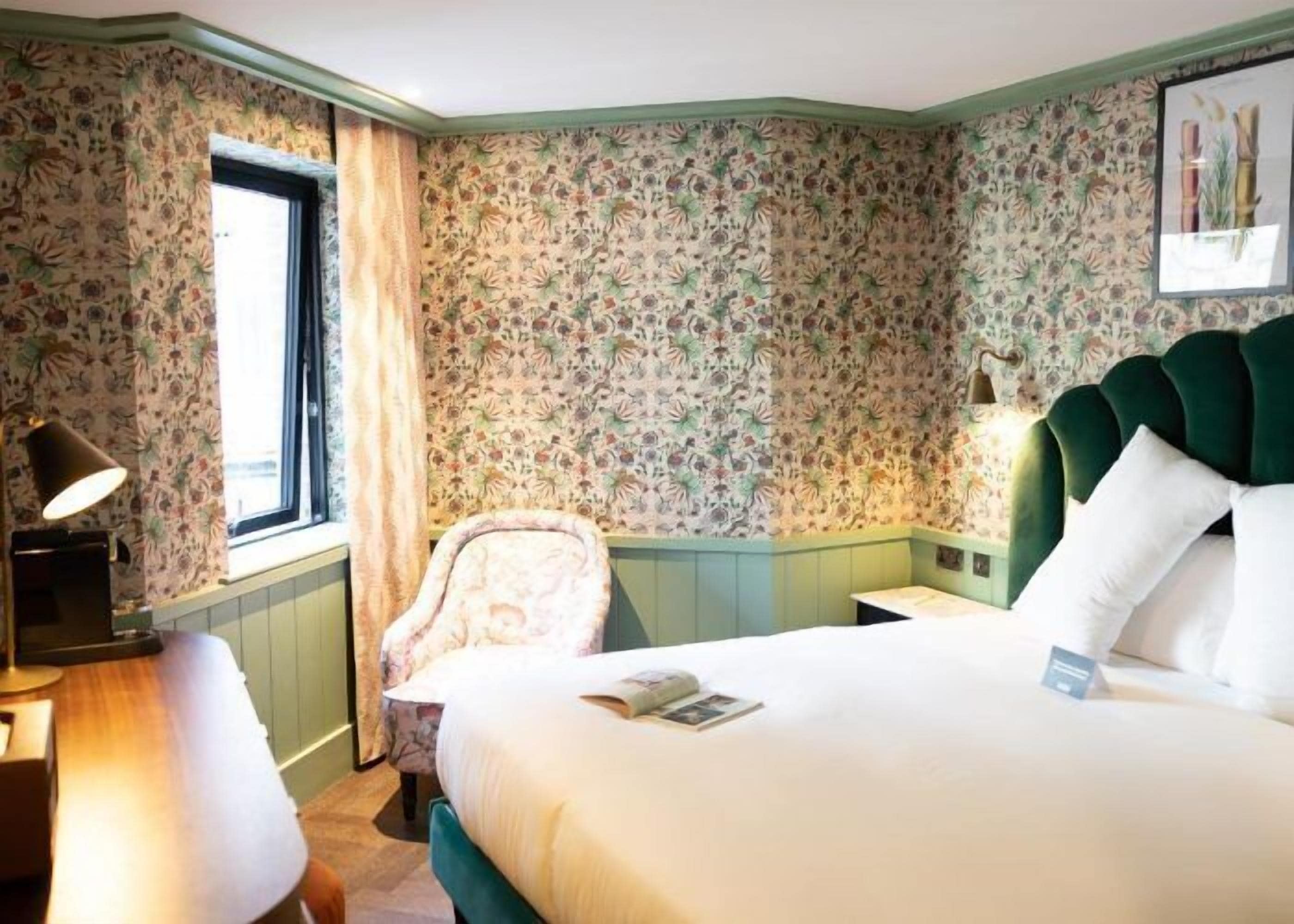 A hotel room with floral wallpaper, a green upholstered headboard, a bedside table with a lamp, a chair, and a bed with white bedding.