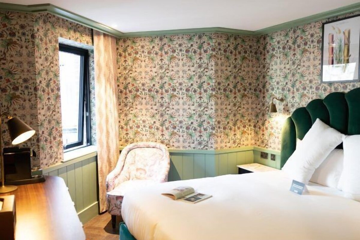 A hotel room with floral wallpaper, a green upholstered headboard, a bedside table with a lamp, a chair, and a bed with white bedding.