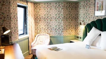 A hotel room with floral wallpaper, a green upholstered headboard, a bedside table with a lamp, a chair, and a bed with white bedding.