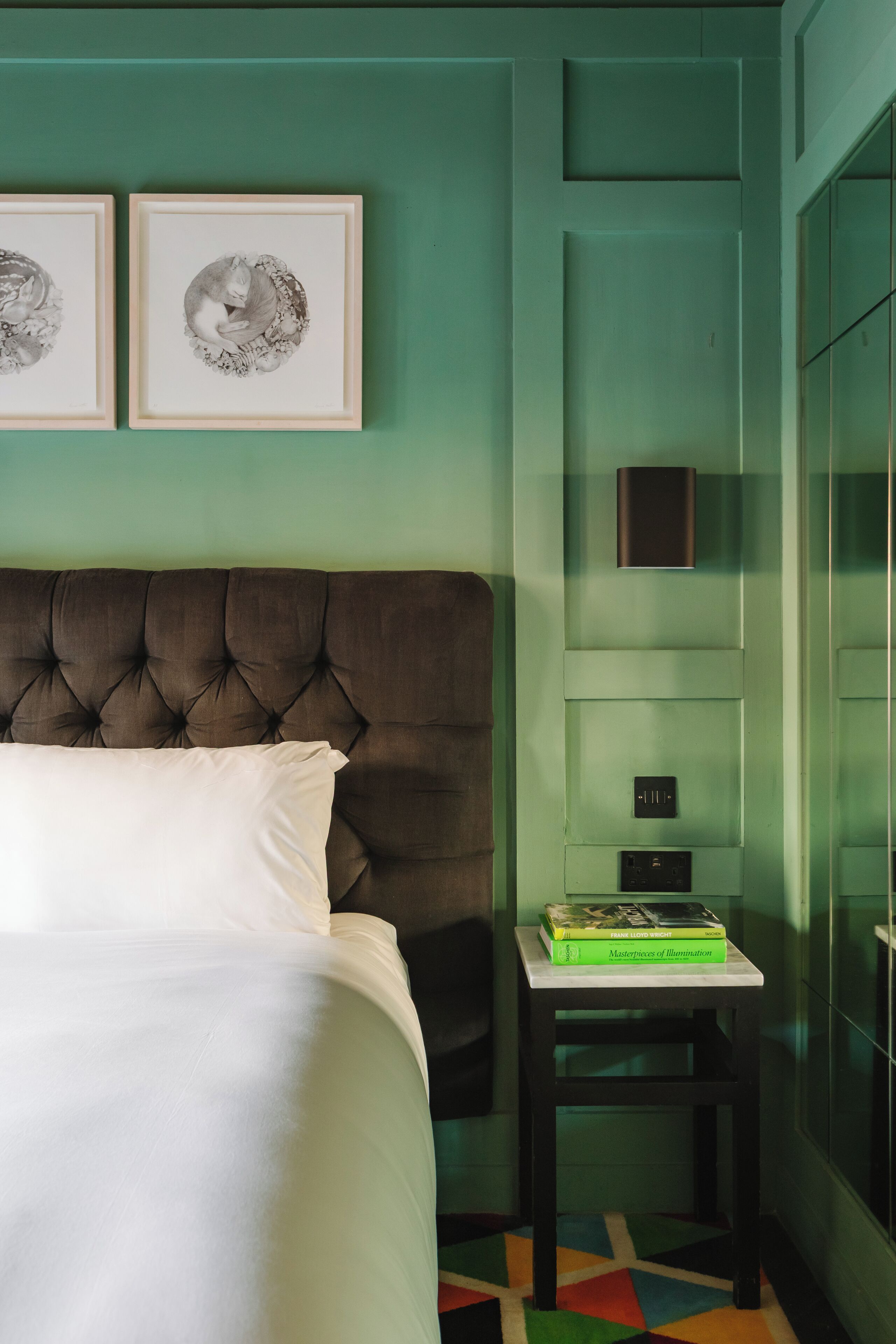 A bedroom with a tufted headboard, a bed with white linens, a small bedside table with a book, and a colorful geometric rug.