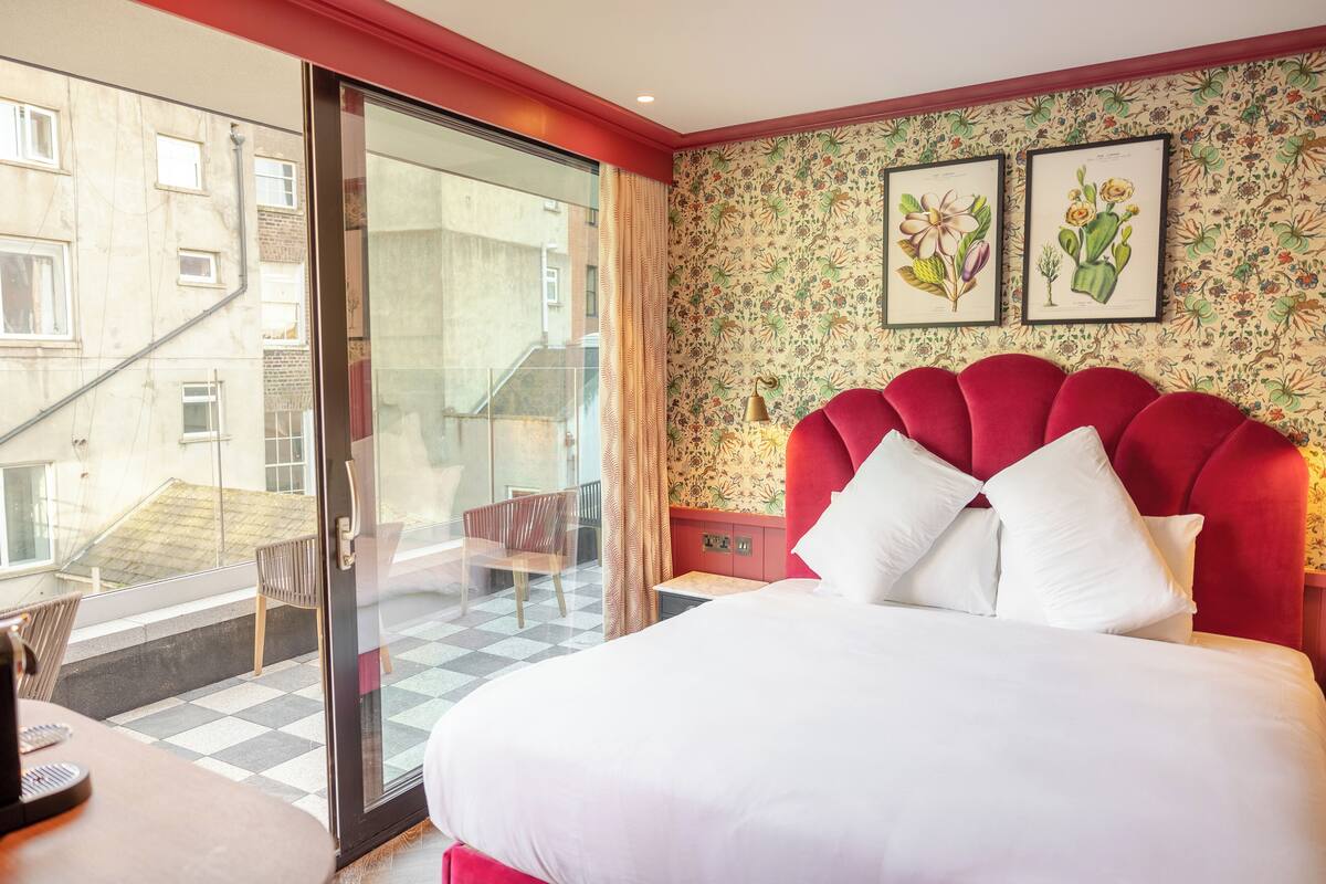 A hotel room with a large bed, a red headboard, a balcony with a checkered floor, and floral wallpaper.