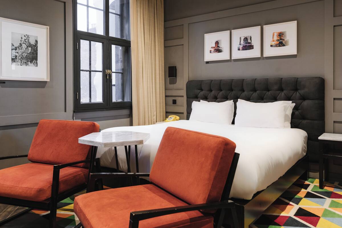 A hotel room with a large bed, two orange chairs, a small table, and artwork on the walls.