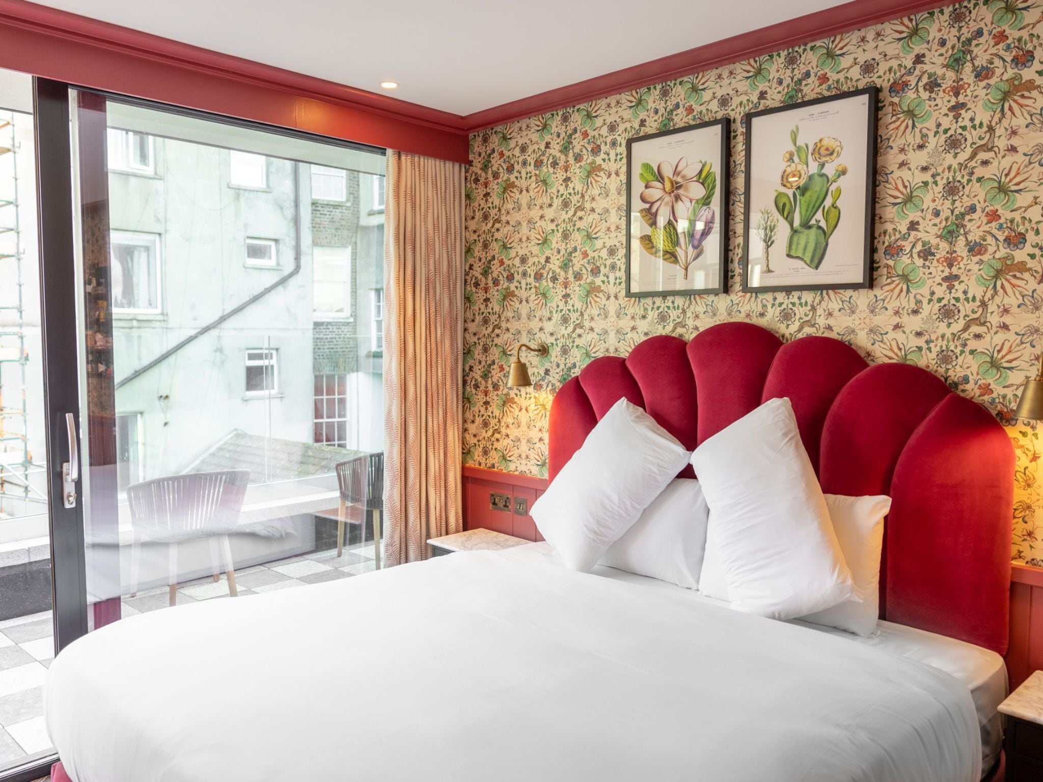 A hotel room with a bed, a window with a view of buildings, a red headboard, and floral wallpaper.
