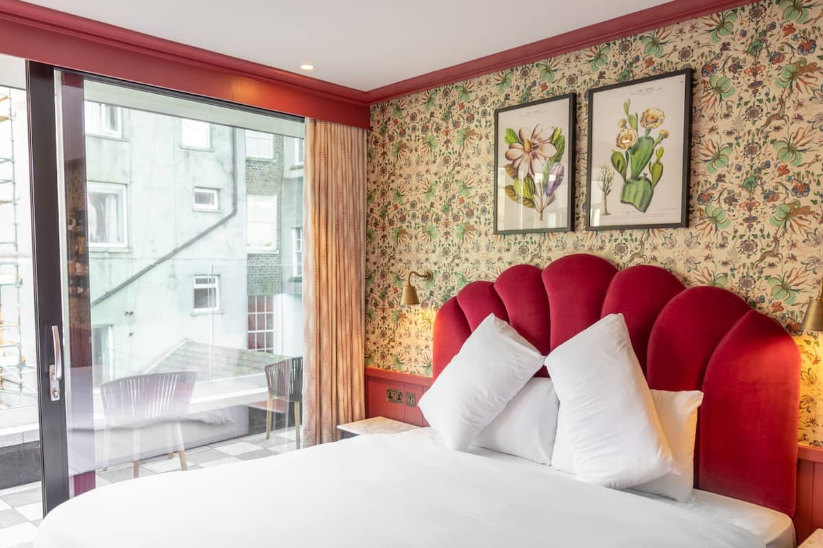 Una habitación de hotel con cama, ventana y vistas a edificios, cabecero rojo y papel tapiz floral.