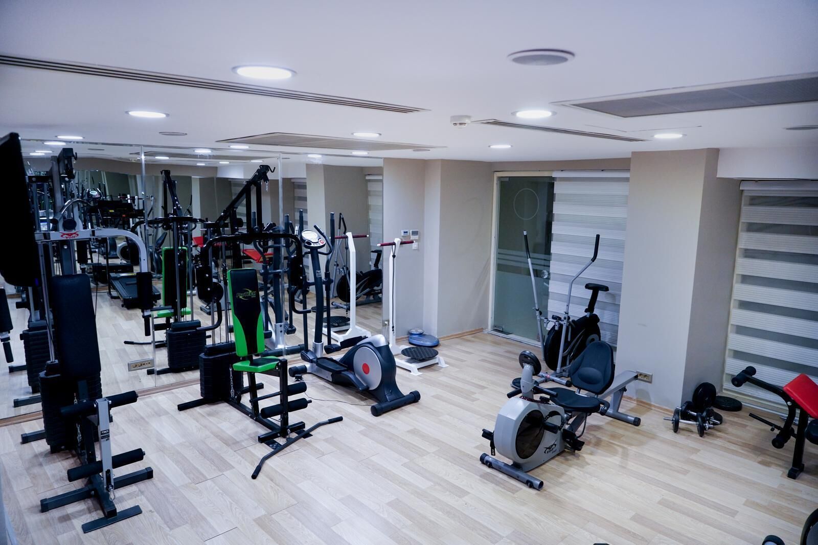 A gym with various exercise machines, a mirrored wall, and a window with blinds.