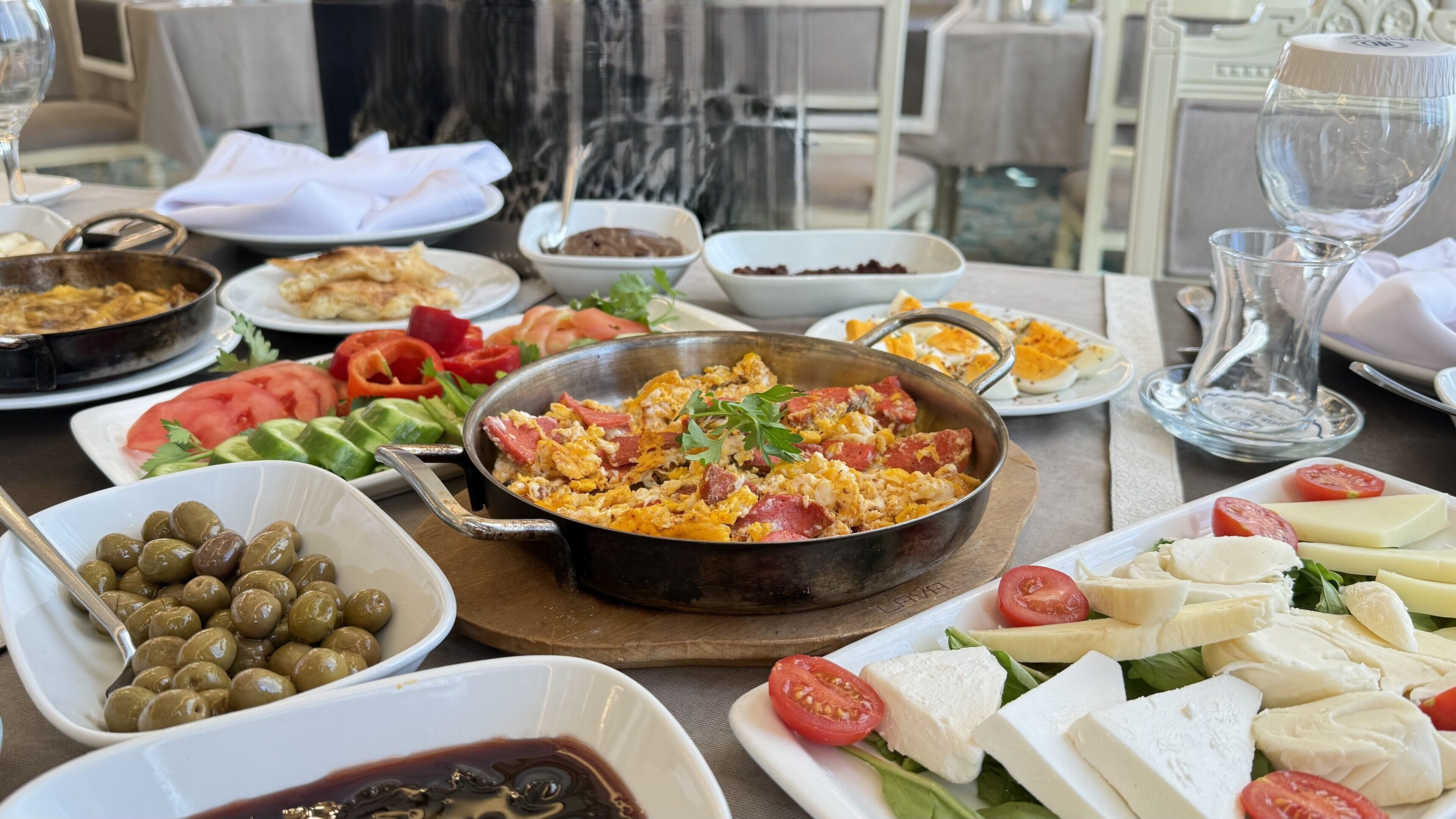 A table set with various dishes including a skillet of scrambled eggs, a bowl of olives, and plates of cheese and tomatoes.