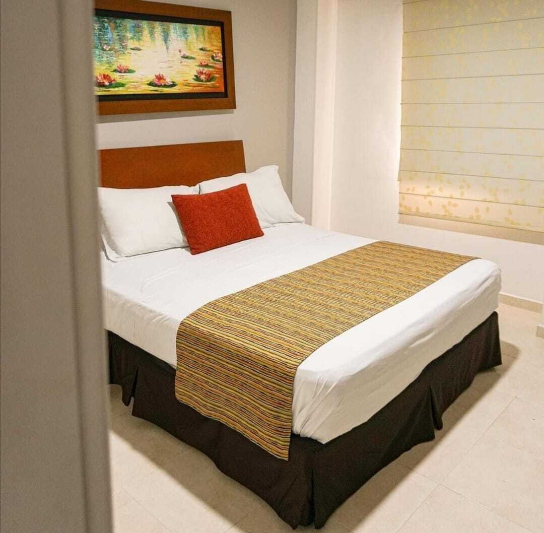 Una cama bien hecha con sábanas blancas y un plaid marrón a rayas, un almohadón rojo y un cabecero de madera.
