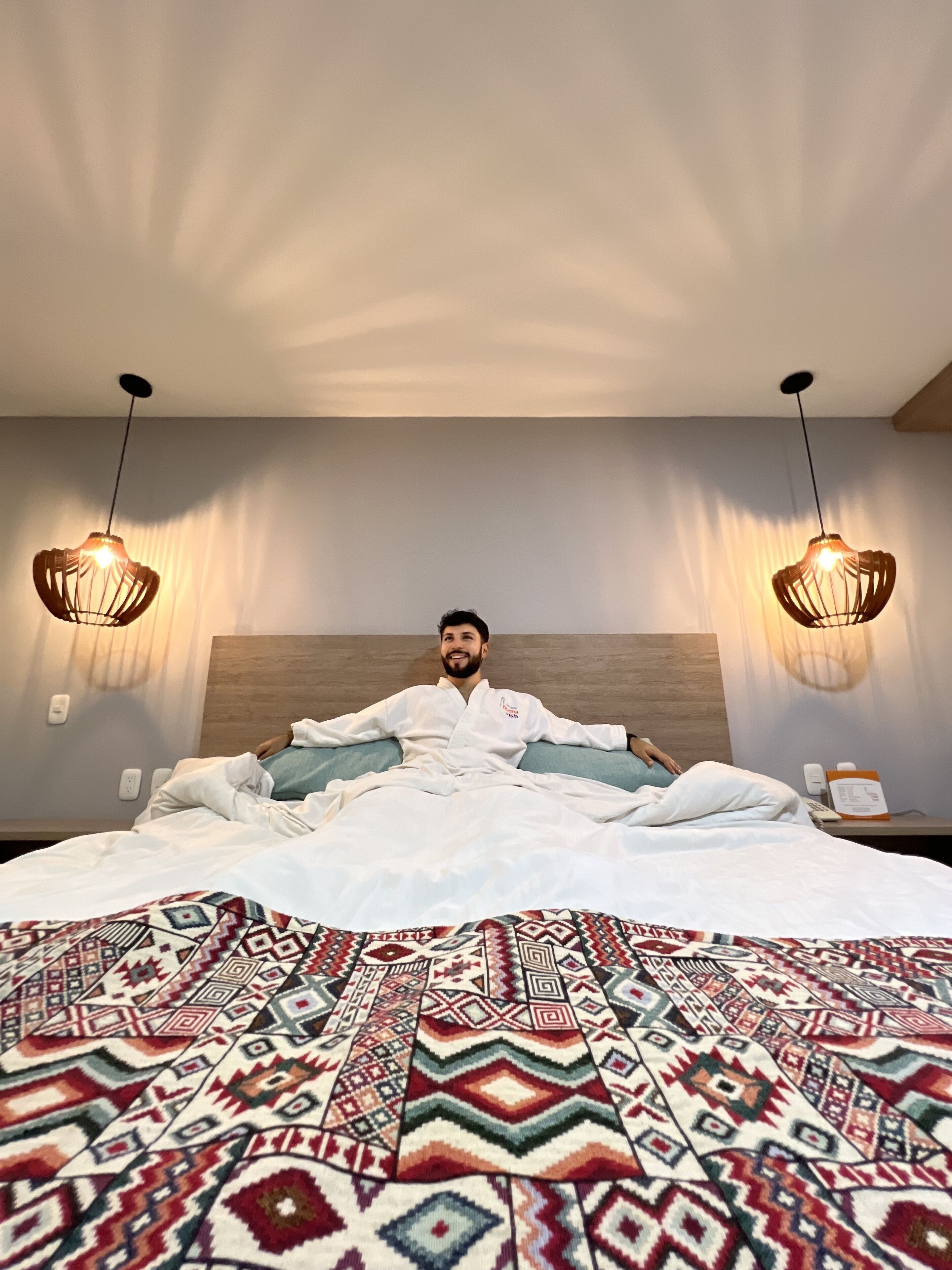 Une chambre d’hôtel avec un grand lit, une couvre-lit à motifs, deux lampes fixées au mur et une tête de lit en bois.