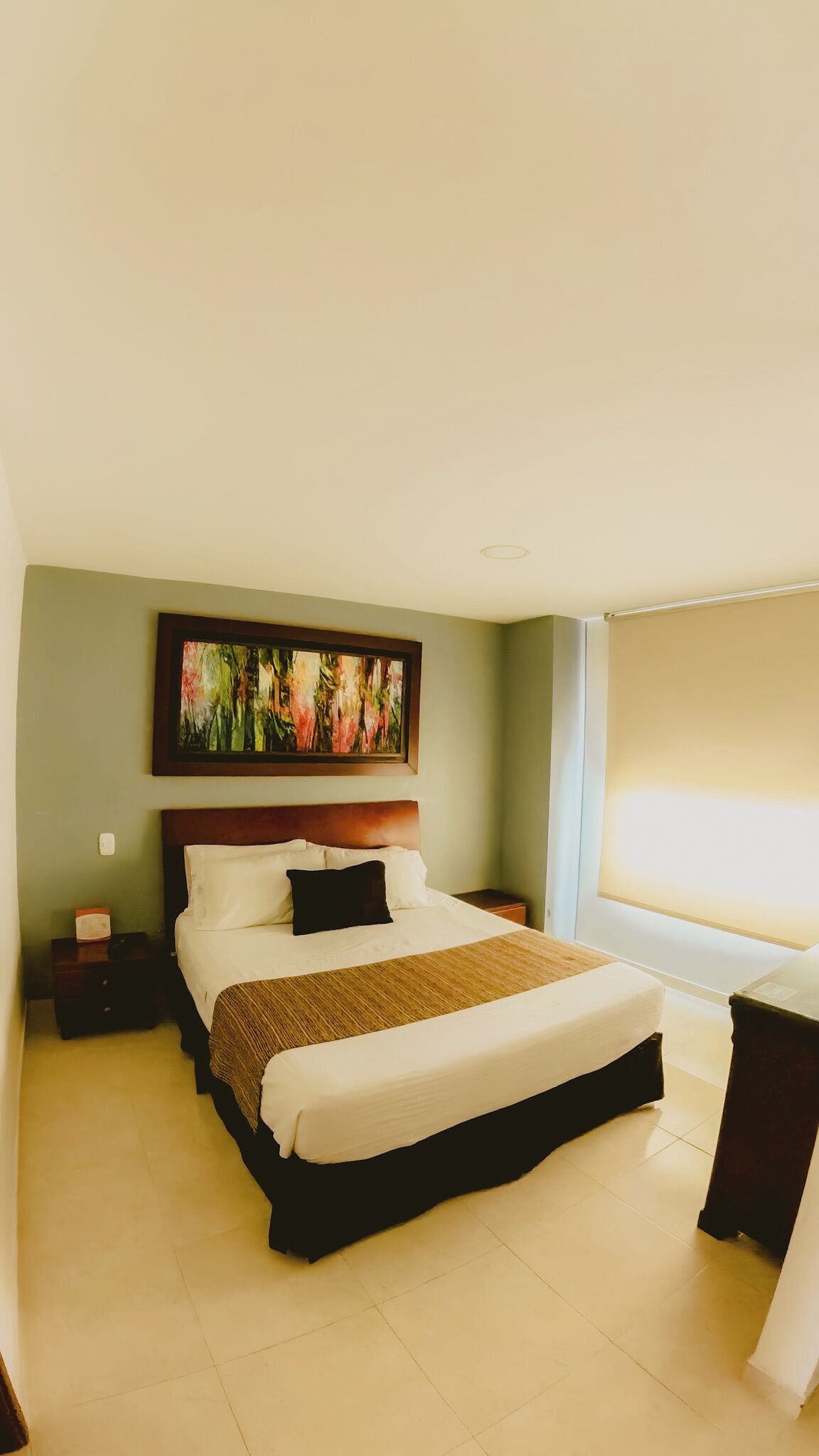 A hotel room with a large bed, a nightstand, a bedside table, a framed abstract painting, and a window with curtains.