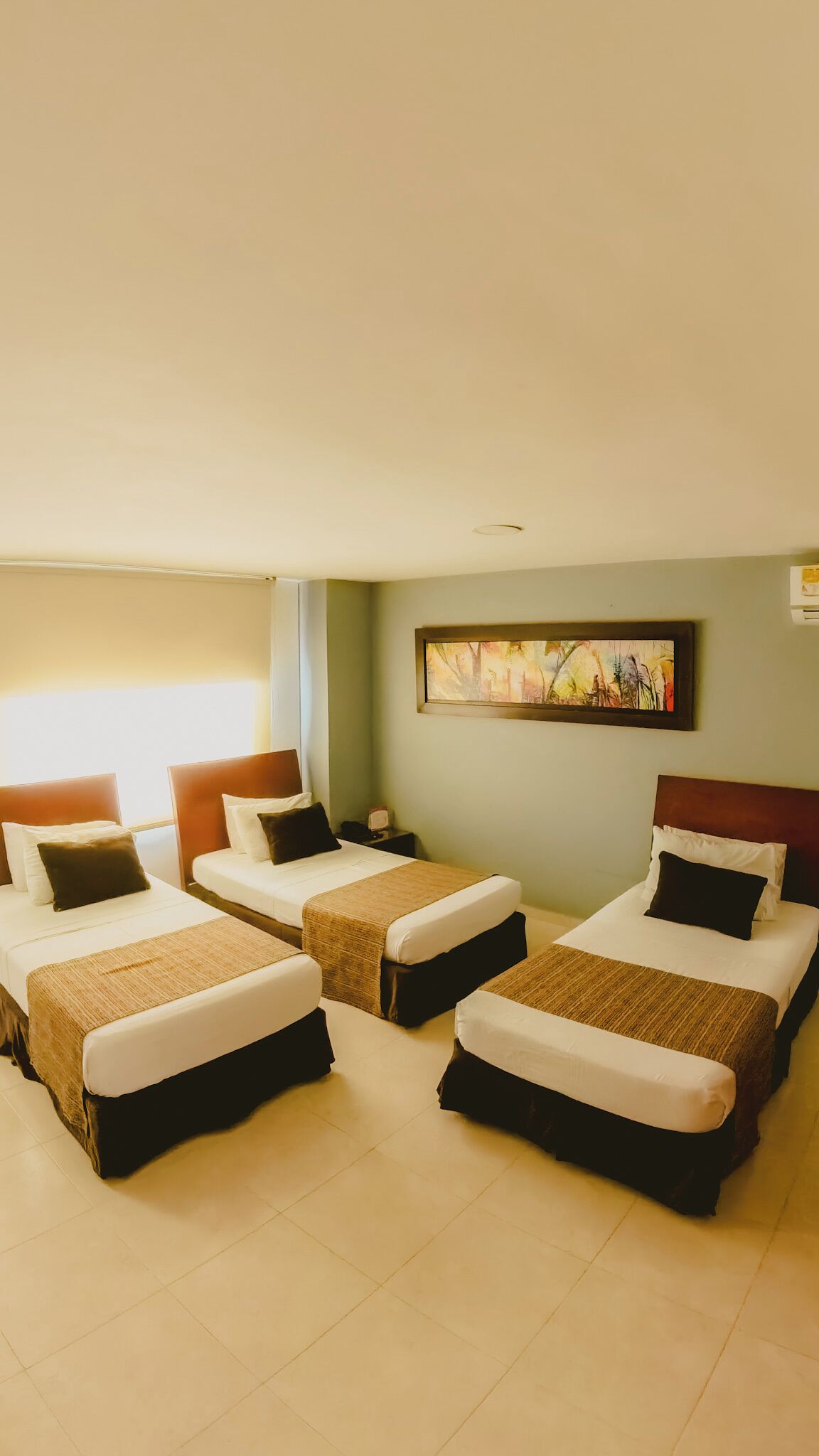 A hotel room with four beds arranged in a U-shape, a painting on the wall, and a nightstand with a lamp.