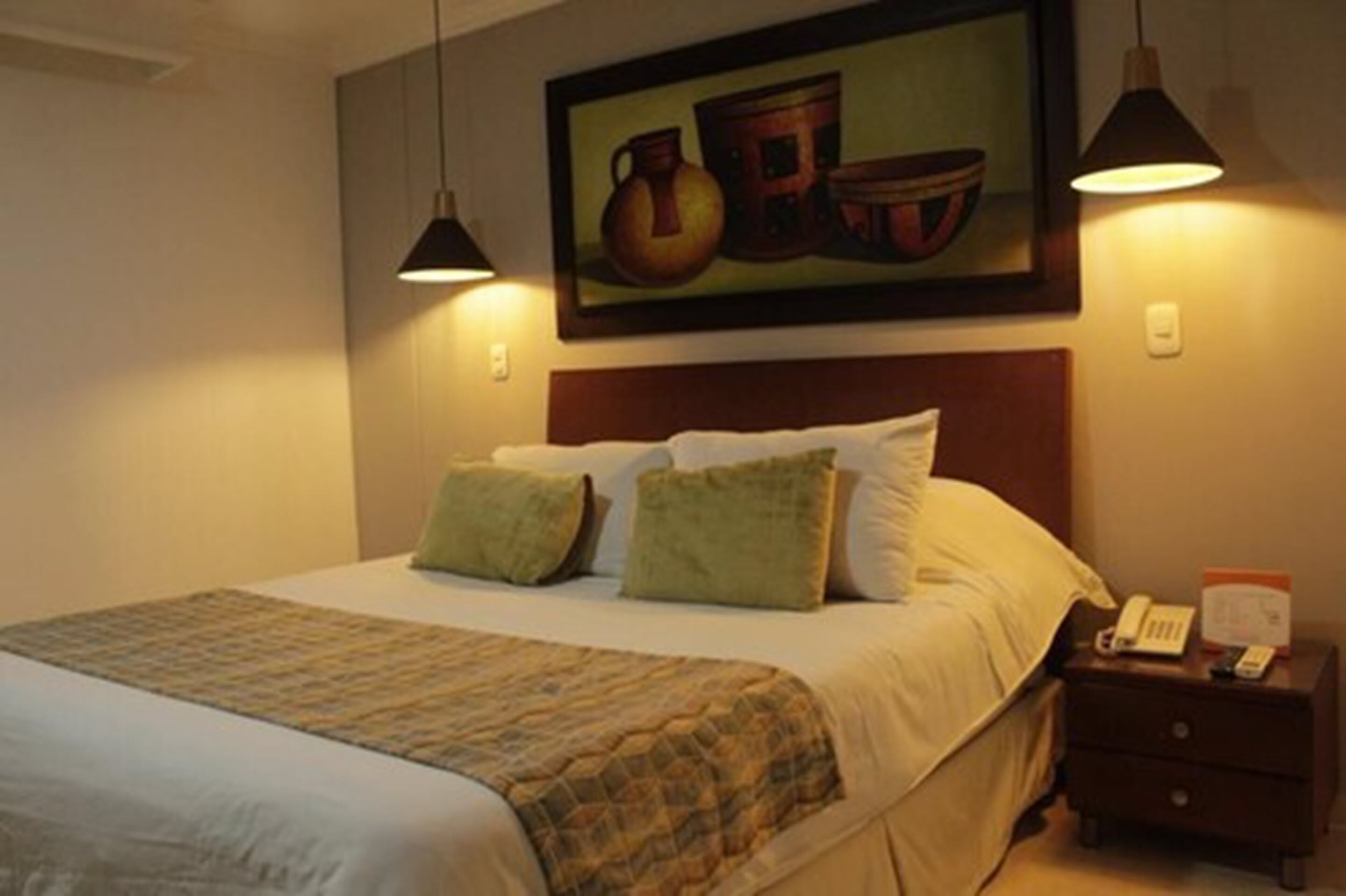 Una habitación de hotel con una cama, una mesita de noche, un cuadro y dos lámparas colgantes.
