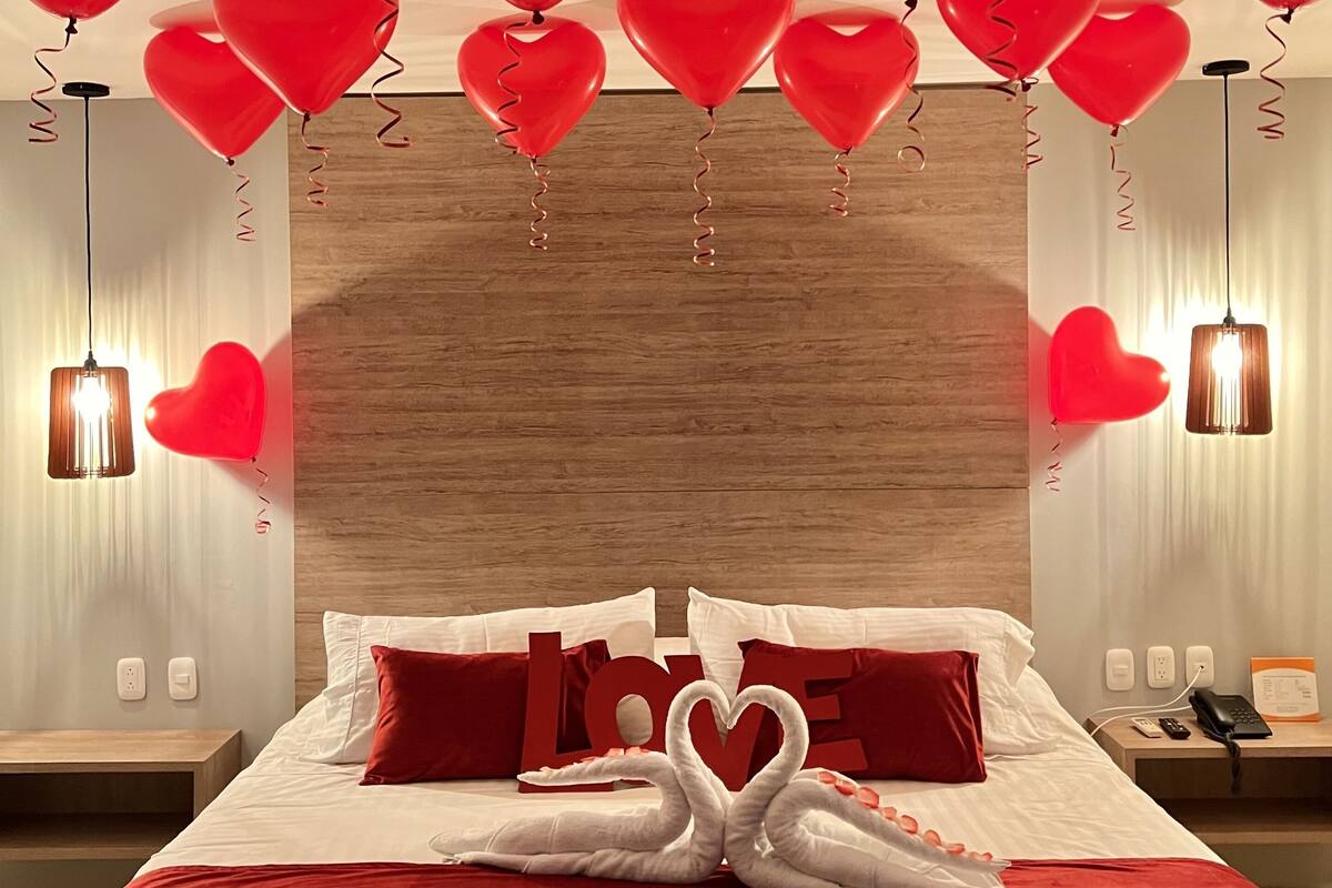 Una habitación de hotel con cabecera de madera, cama con ropa de cama roja y blanca, globos con forma de corazón y una decoración de cisne.