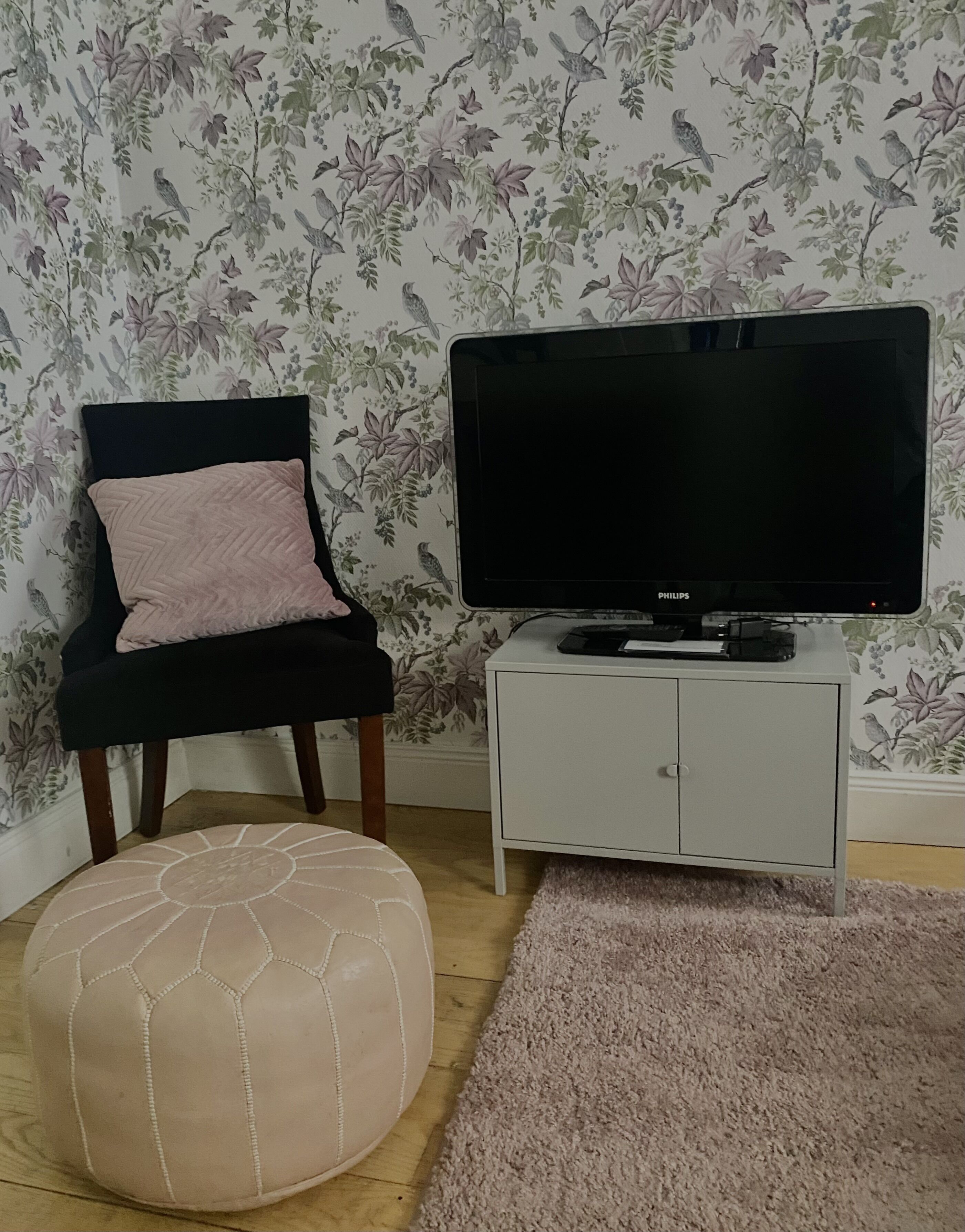 A room with a floral wallpaper, a black chair with a pink cushion, a television on a white cabinet, and a beige ottoman on the floor.