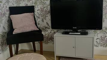 A room with a floral wallpaper, a black chair with a pink cushion, a television on a white cabinet, and a beige ottoman on the floor.
