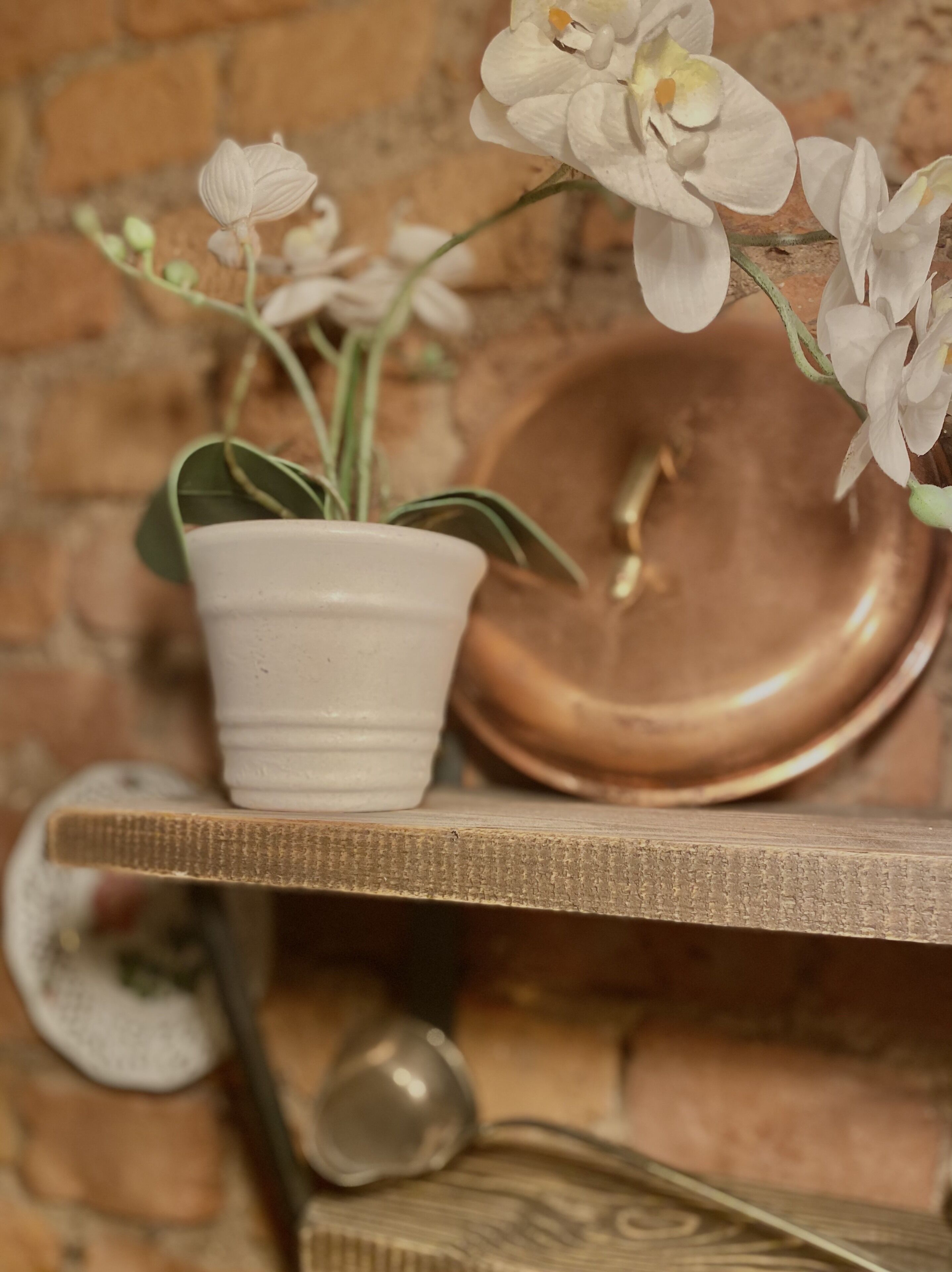 Een witte orchidee in een pot op een plank met een koperen kom en een kleine metalen lepel.