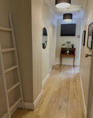 A hallway with wooden flooring, a white wall, a wooden console table with plants, a mirror, and a door with a brass handle. - Lilla Brunn - Hostel (Stockholm)