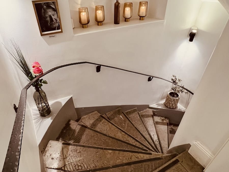 Una escalera con barandilla de madera, un jarrón con una flor rosa e iluminación empotrada en la pared.