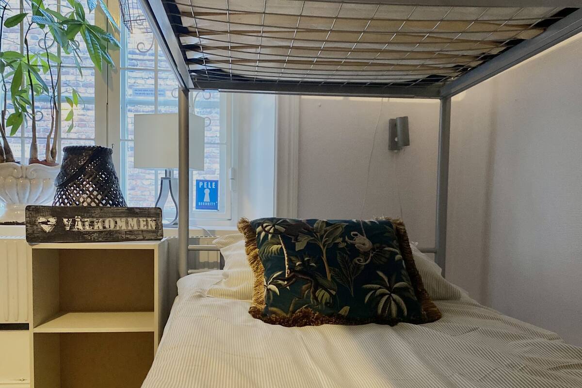 Un dormitorio con cama, mesita de noche, planta en maceta y ventana con vistas al exterior.