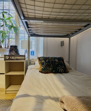 A bedroom with a bed, a nightstand, a potted plant, and a window with a view of the outside. - Lilla Brunn - Hostel (Stockholm)