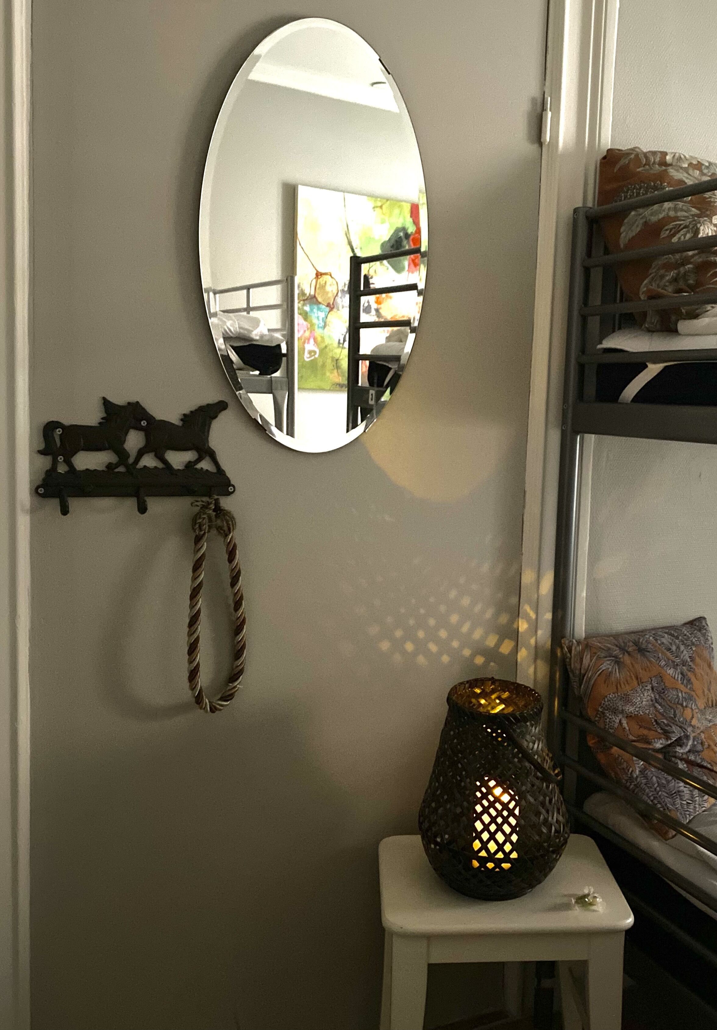 Um quarto com um espelho redondo na parede, uma pequena mesa branca com um abajur decorativo e uma estante com almofadas.